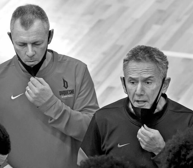 Duquesne head mens basketball coach Keith Dambrot (right) flanked by assistant coach Terry Weigand pull down their face cover during a timeout during a game.